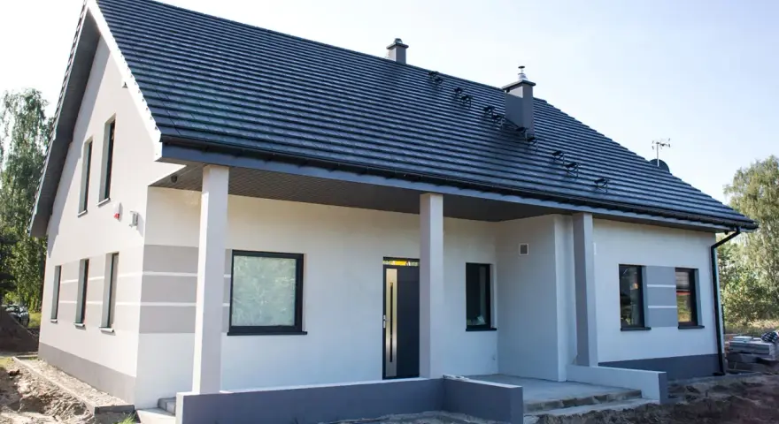 Nowy dom z szarą dachówką i jasną elewacją, ozdobioną subtelnymi, poziomymi pasami. Trwają prace nad malowaniem elewacji, nadając budynkowi nowoczesny wygląd.