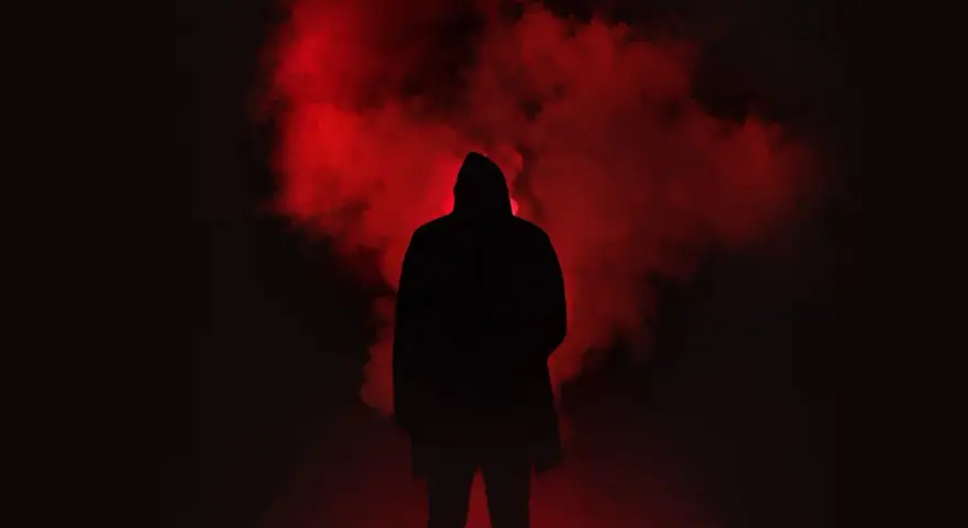 Figura incappucciata nell'oscurità, circondata da fumo rosso. Un'immagine che evoca la differenza tra giallo e noir, tra mistero e oscurità.
