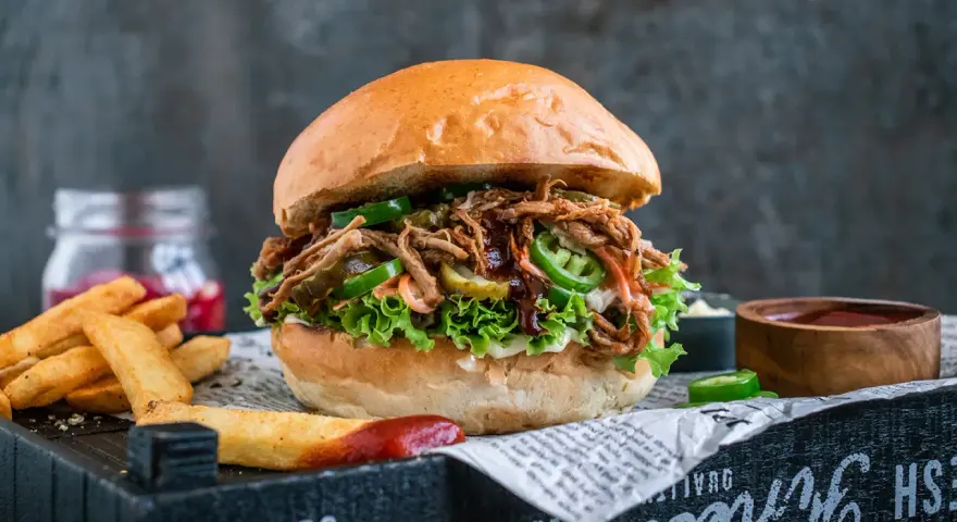 Saftiger pulled pork burger mit Salat, Jalapeños und Gurken. Die Zutaten für diesen Burger sind perfekt aufeinander abgestimmt. Dazu Pommes und Dip.