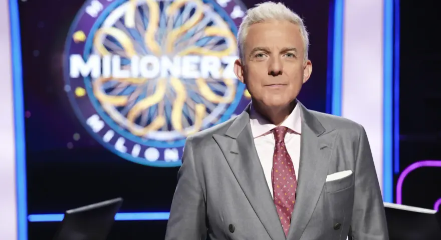 Hubert Urbański w garniturze, na tle koła z napisem "Milionerzy". Co dajemy komuś na drogę milionerzy? Wiedzę i odwagę!
