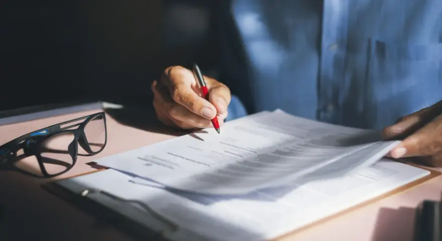 Persona revisa y firma una nota de encargo inmobiliaria, con gafas y bolígrafo rojo sobre documentos.