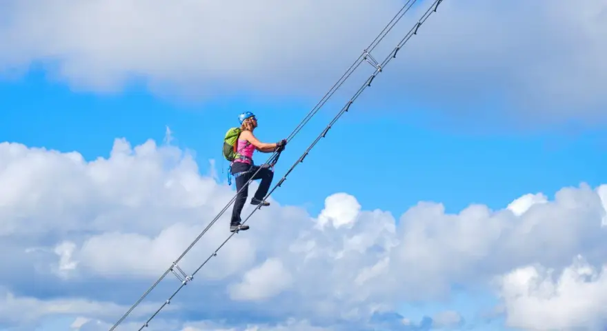 Frau klettert auf dem Donnerkogel Klettersteig über eine Hängebrücke, umgeben von blauem Himmel und Wolken.