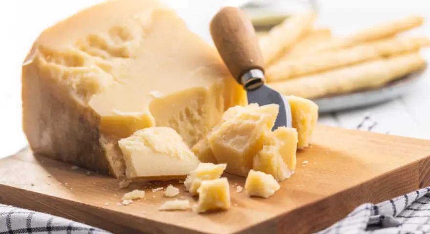 Ein Stück Parmesan mit grob abgebrochenen Stücken und einem Käsemesser. Die Parmesan Rinde ist sichtbar.