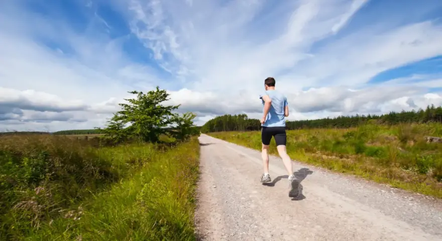Uomo corre su strada sterrata, ideale per la preparazione 10 km principianti. Cielo azzurro con nuvole bianche.