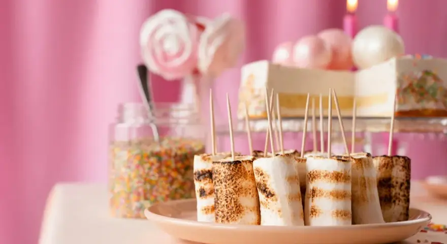 Rostade marshmallows på spett, perfekt för roliga recept att baka till kalaset. En burk strössel och en tårta i bakgrunden.