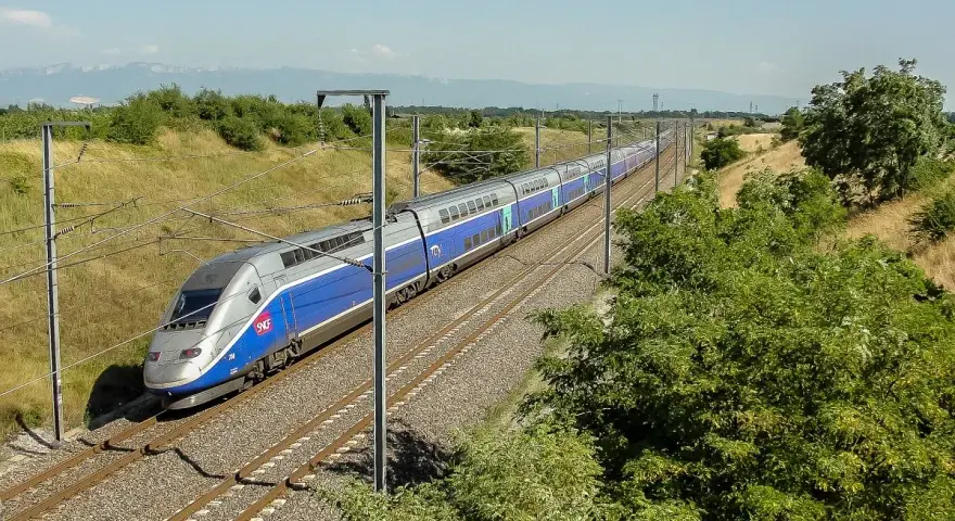 Niebiesko-srebrny pociąg dużych prędkości TGV na torach kolejowych, otoczony zielenią i słupami trakcyjnymi.