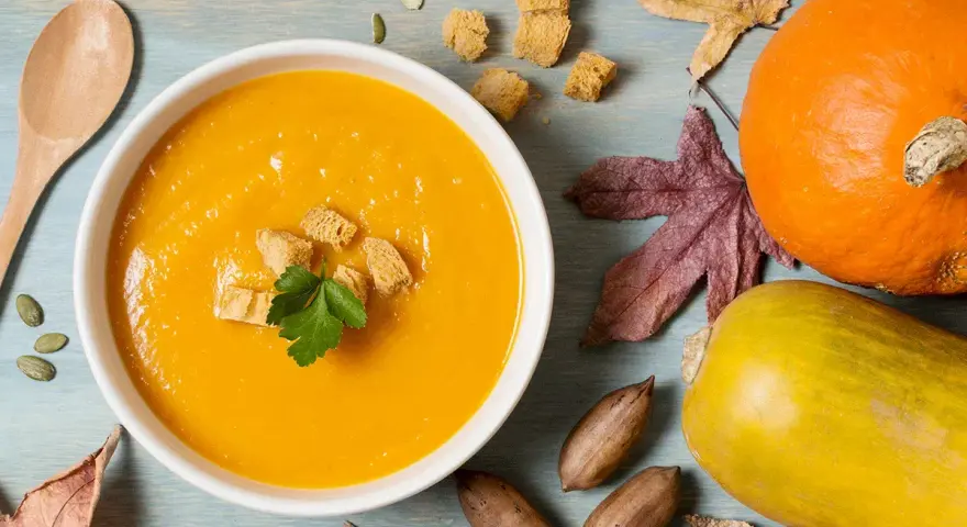 Un reconfortante tazón de puré de calabaza, adornado con picatostes y perejil, junto a calabazas, hojas de otoño y nueces.