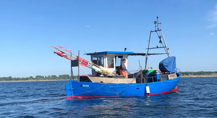 Blaues Fischerboot "RERIK" mit roten Markierungen auf dem Wasser. Ein Mann steht an Deck, rote Fahnen ragen empor.