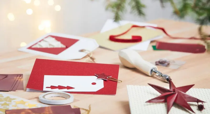 Förberedelser för att måla julkort. Rött glitterpapper, vita etiketter med julgransmotiv och en stjärnformad gemklämma ligger på ett träbord.