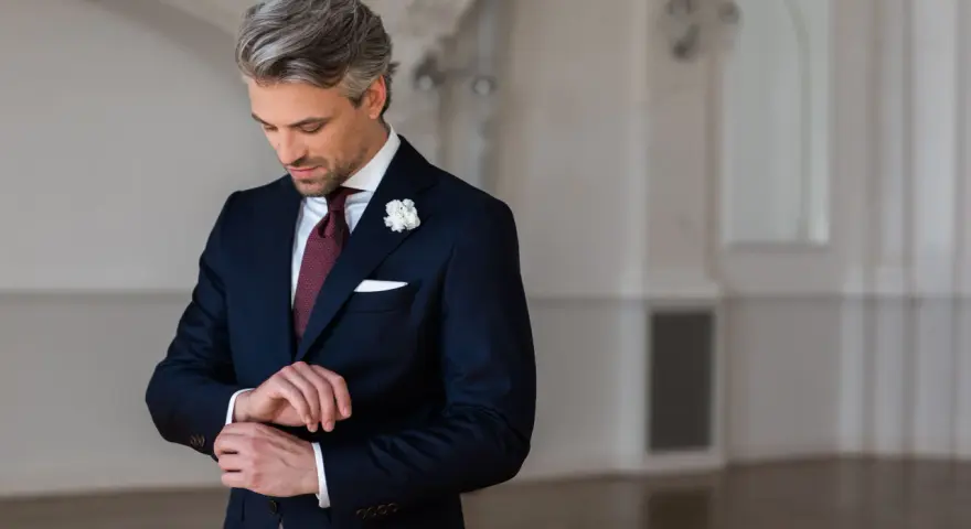 Elegancki mężczyzna w granatowym garniturze i bordowym krawacie poprawia mankiet.