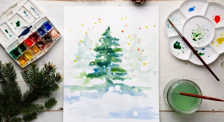 Pintura de acuarela de un árbol de Navidad nevado, con una paleta de acuarelas y pinceles, ideal para una acuarela navidad facil.
