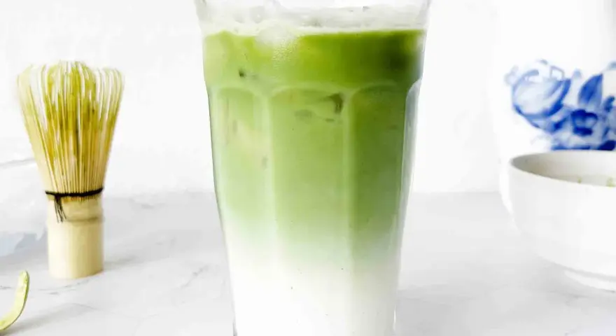 En isig matcha latte med lager av mjölk och grönt te. Bredvid står en bambuvisp, redo att blanda nästa goda dryck.