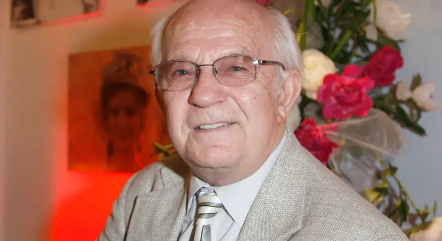 Uśmiechnięty Jerzy Antczak, reżyser znany z niezapomnianych filmów, w eleganckim garniturze, na tle bukietu kwiatów i portretu.