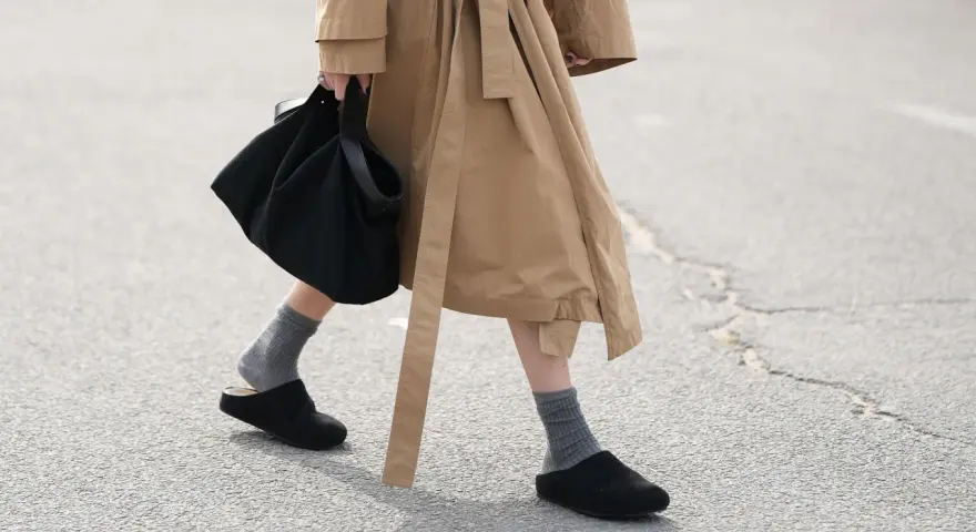 Persona camina con zuecos negros y calcetines grises, llevando un bolso negro y un abrigo beige.