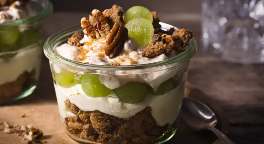 Ein Glas mit cremigem Dessert, geschichtet mit grünen Trauben, zerbröselten Keksen und Nüssen. Ein köstliches Trauben-Cookie-Dessert, perfekt für jeden Anlass.