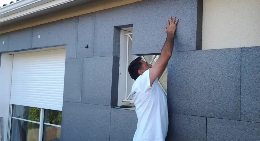 Un ouvrier pose des panneaux isolants gris pour l'isolation par l'extérieur d'une maison, près d'une fenêtre avec volet roulant.