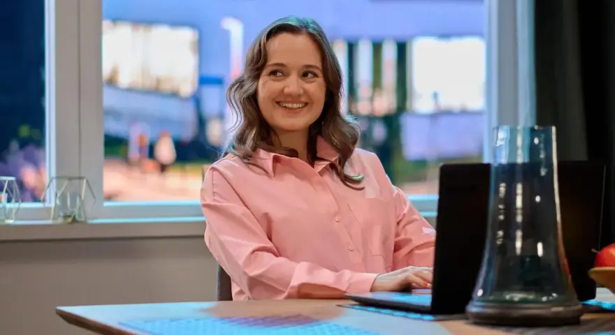Uśmiechnięta Dorota Krempa pracuje przy laptopie, siedząc przy stole. W tle widać okno z nocnym widokiem.