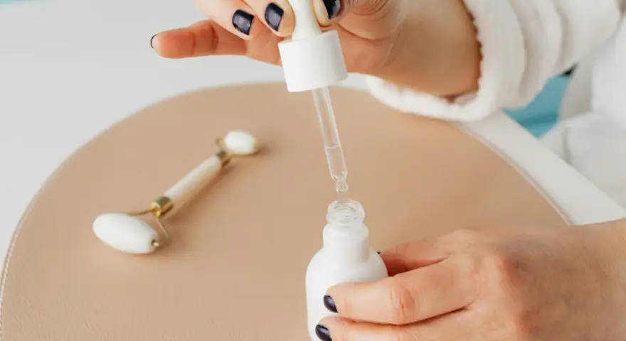 Dłoń z ciemnym lakierem do paznokci aplikuje serum z pipety do białej buteleczki. Obok leży jadeitowy roller. Zastanawiasz się, z czym łączyć niacynamid?