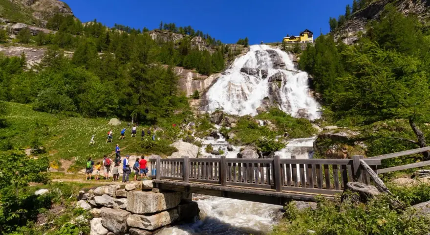 Cascata imponente con escursionisti su un sentiero, un ponte di legno attraversa un torrente. Un esempio di cosa vedere in Piemonte per gli amanti della natura.