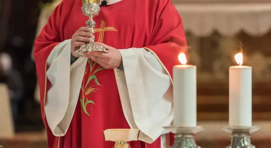 Kapłan w czerwonej szacie z krzyżem trzyma kielich podczas konsekracji. Płoną dwie świece.