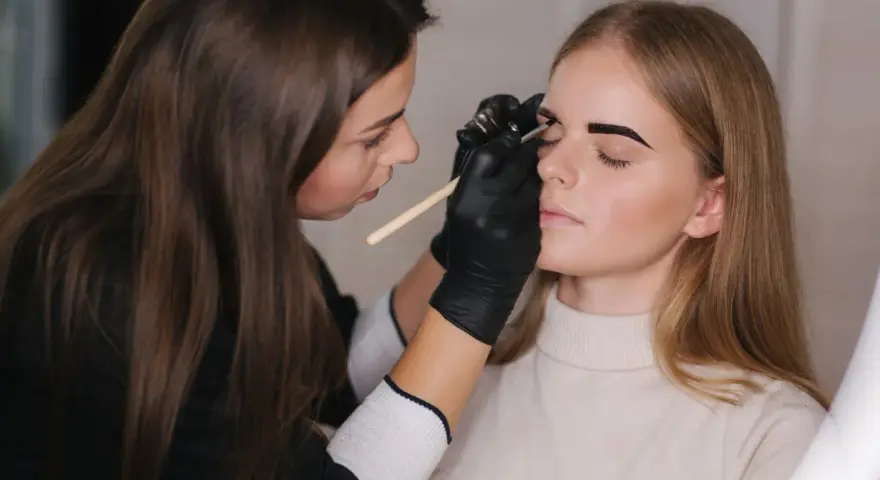 Stylistka w czarnych rękawiczkach precyzyjnie maluje brwi klientce. Czy to henna czy farbka, efekt będzie zachwycający.