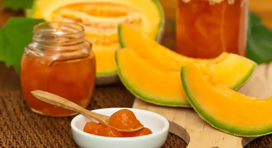 Marmellata di melone in vasetti, con fette di melone fresco e un cucchiaino pieno di confettura.
