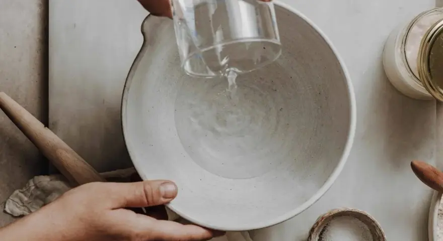 Wasser wird in eine Schüssel gegossen, vielleicht zu viel Wasser im Brotteig. Ein Nudelholz liegt daneben.
