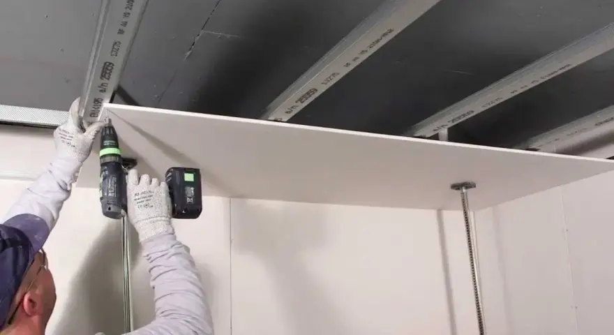 Persona con guanti avvita una lastra di cartongesso al soffitto metallico, un progetto di cartongesso fai da te.
