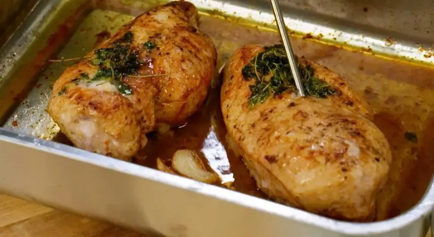 Två saftiga, gyllene grillade kycklingbröst i en ugnsform med örter och vitlök. En termometer mäter temperaturen på den ena kycklingen.
