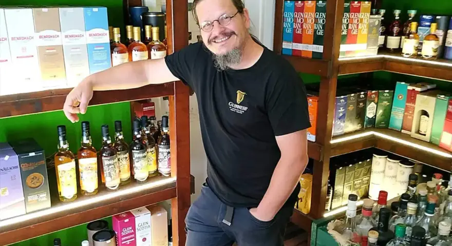 Mann in einem Guinness-T-Shirt steht in einem Laden voller Whisky-Flaschen und -Schachteln. Er lächelt und lehnt sich an ein Regal.