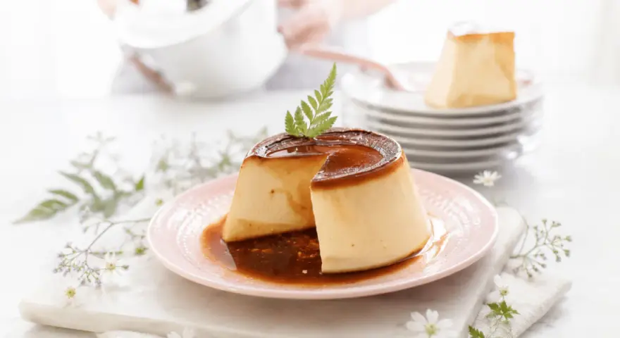 Delicioso flan de queso con caramelo líquido, decorado con una hoja verde. En el fondo, una persona prepara más postres.