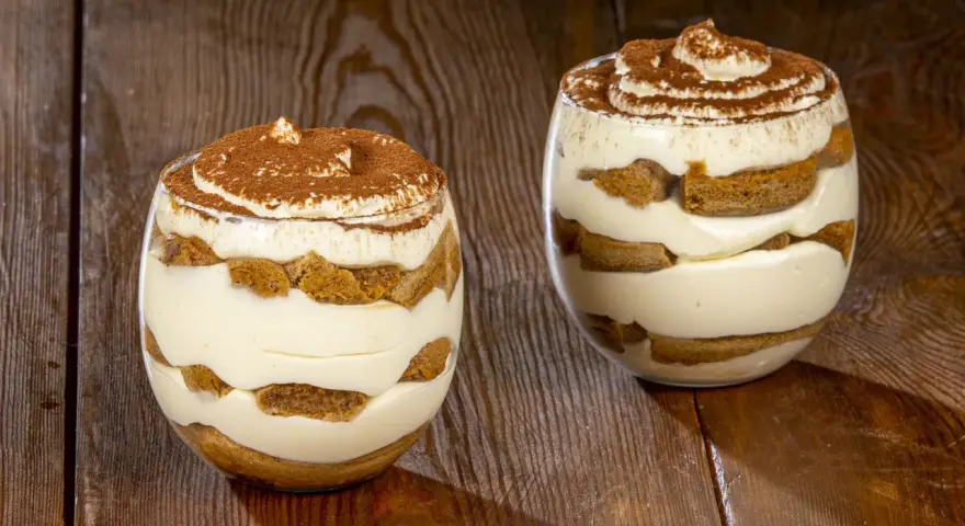 Due bicchieri di tiramisù senza uova, stratificati con crema e savoiardi, spolverati di cacao.