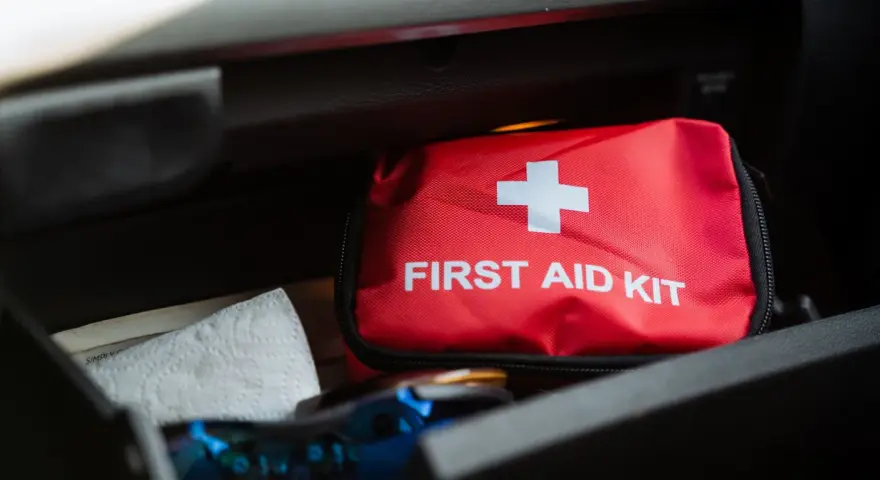 Czerwona apteczka samochodowa z białym krzyżem i napisem "FIRST AID KIT" leży w schowku. Obok niej papierowy ręcznik.