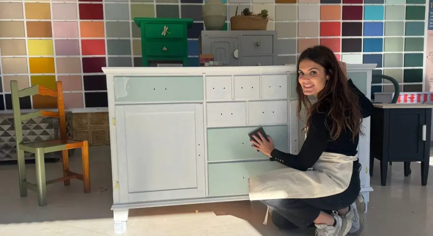 Mujer sonríe mientras lija un mueble para pintar. Detrás, un muro con muestras de colores de pintura.
