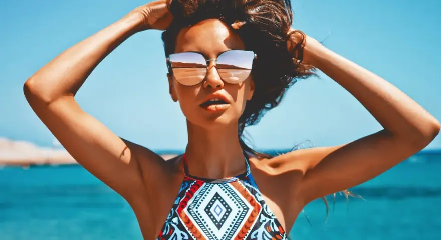Kobieta w okularach przeciwsłonecznych na plaży, z rękami w gorze. Jej skóra ma piękny, złocisty odcień, jakby świadczył o wspaniałych beta karoten efektach.