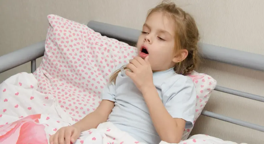 Mała dziewczynka z **przewlekłym kaszlem u dziecka** leży w łóżku, trzymając termometr.