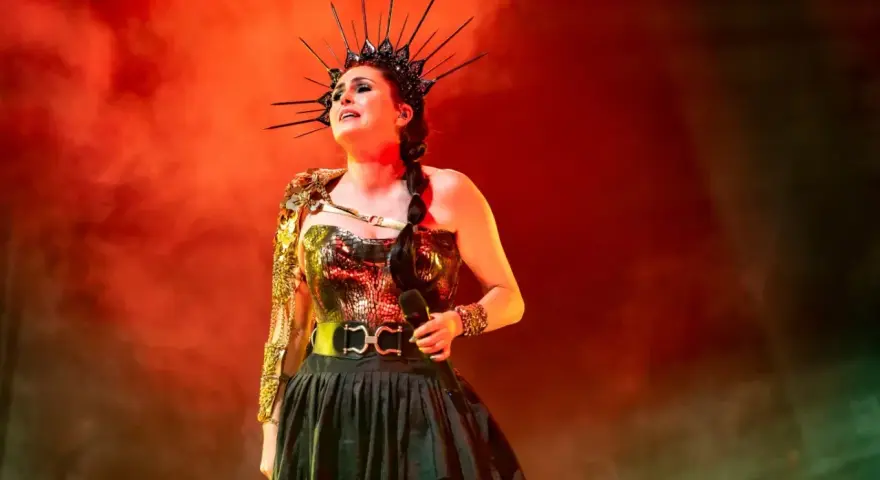 Wokalistka Within Temptation w koronie i błyszczącej kreacji, z mikrofonem w dłoni, na tle czerwonej mgły.