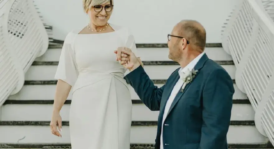 Elegancka kobieta po pięćdziesiątce w sukience do ślubu cywilnego, trzymająca za rękę pana młodego w granatowym garniturze.