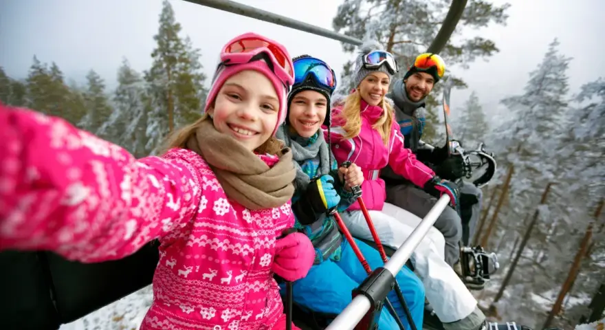 Rodzina na wyciągu narciarskim, robiąca selfie. Dziewczynka z przodu uśmiecha się szeroko. To ich wymarzone wczasy pod gruszą, ale zimowe!