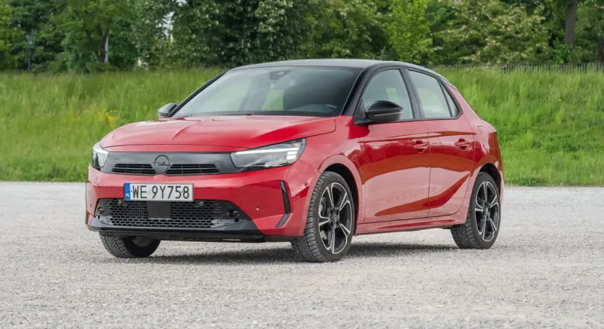 Czerwony Opel Corsa E na tle zieleni. Zbliżenie na przód auta, gdzie widoczne są światła LED i tablica rejestracyjna. Poznaj opel corsa e wymiary.