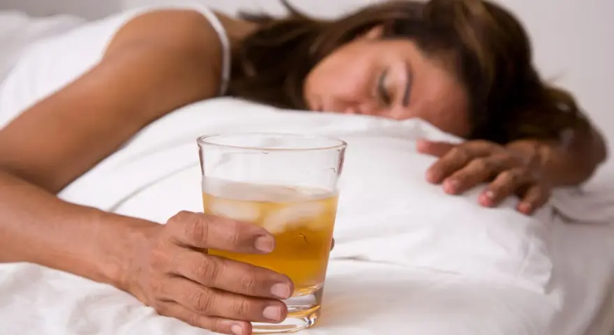 Kobieta w łóżku z drinkiem, nieświadoma. Objawy po zażyciu pigulka gwaltu mogą być różne.