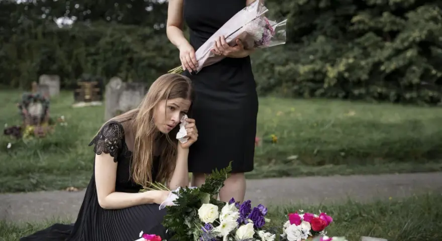 Smutna kobieta w czarnej sukience klęczy przy grobie, ocierając łzy. Obok stoi druga kobieta z bukietem kwiatów. To obraz żałoby, trudny urlop macierzyński po poronieniu.