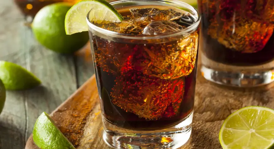 Orzeźwiający Cuba Libre, czyli rum z colą i limonką, z kostkami lodu i świeżymi ćwiartkami cytrusów na drewnianym blacie.