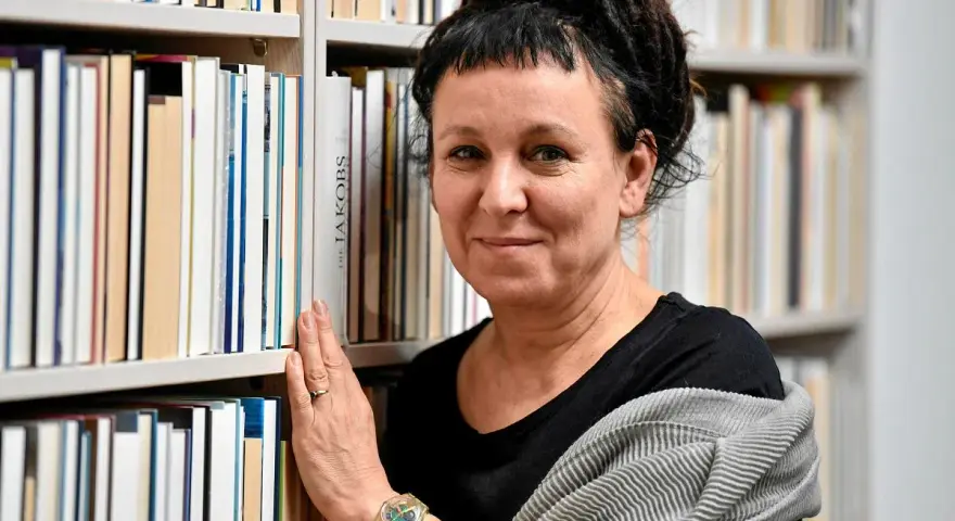 Olga Tokarczuk, w swoim wieku, otoczona książkami, z dłonią na grzbietach.
