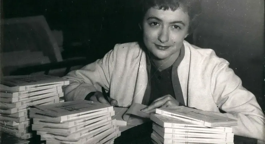 Françoise Sagan vid ett bord med högar av böcker, hon skriver på ett papper.