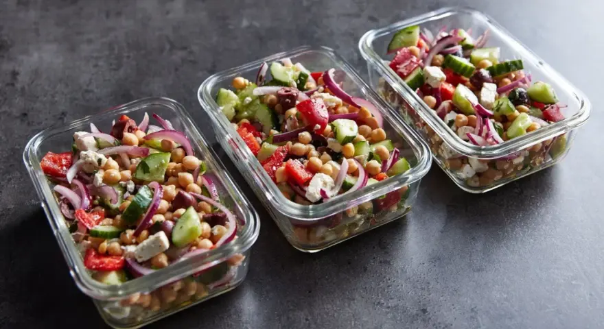 Trzy szklane pojemniki wypełnione kolorową sałatką z ciecierzycą, ogórkiem, pomidorem, czerwoną cebulą i fetą. Idealna na lunch.