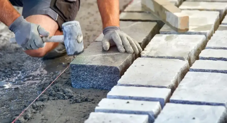 Ręce w rękawiczkach układają kamienne kostki na podsypce. Widać młotek i sznur geodezyjny. Prawidłowe suchy beton proporcje to klucz do trwałej nawierzchni.