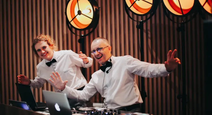 Dwóch DJ-ów w białych koszulach i czarnych muszkach, z uśmiechem na twarzy, przygotowuje się do imprezy. Zastanawiasz się, ile kosztuje DJ na wesele?