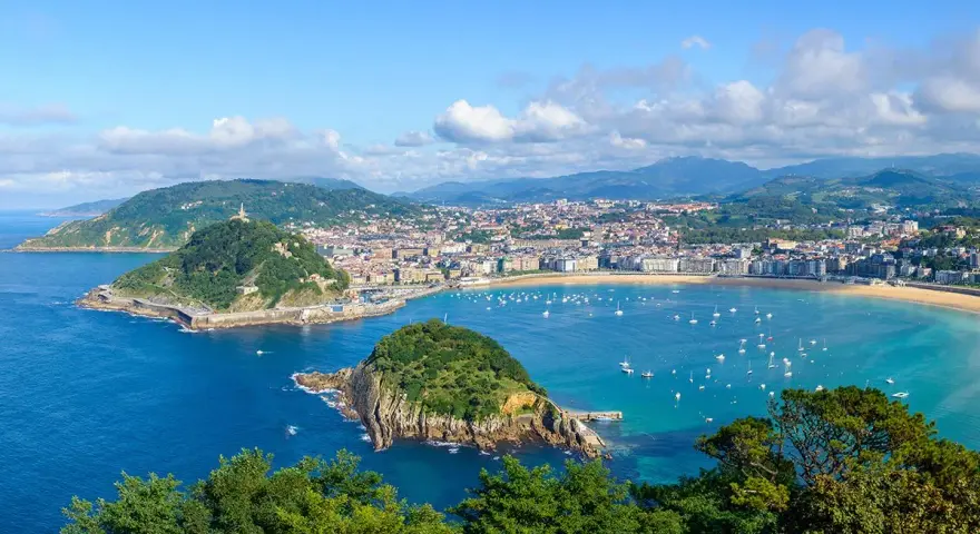 Widok na zatokę La Concha w Donostia San Sebastian, z zielonymi wzgórzami, miastem i licznymi łodziami na turkusowej wodzie.