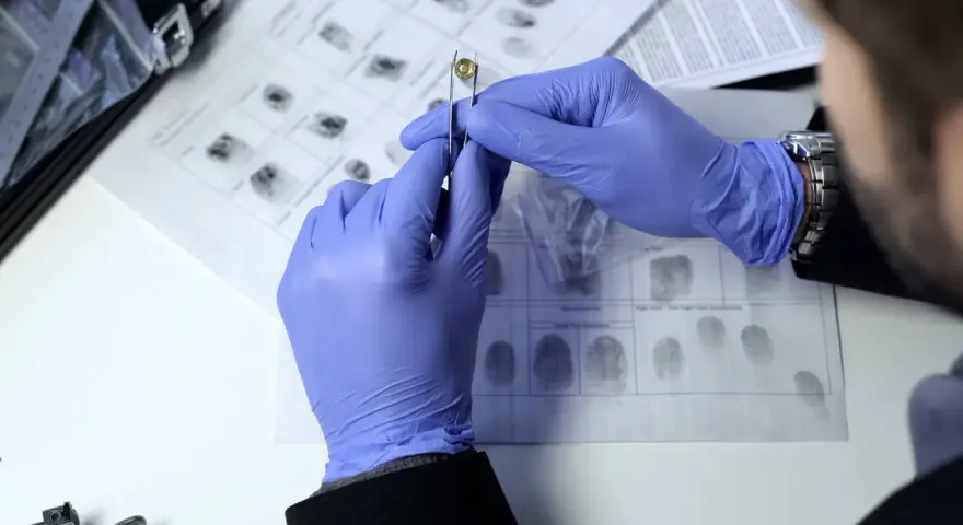 Ein Forensiker untersucht eine Patrone mit einer Pinzette. Im Hintergrund sind Fingerabdrücke zu sehen.